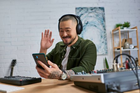 Ein lächelnder asiatischer Mann mit Kopfhörern sitzt an einem Schreibtisch in einem Studio und winkt mit einem Smartphone.