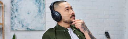 Ein gutaussehender asiatischer Mann mit Kopfhörern in einem Studio-Setting, verloren in Gedanken.