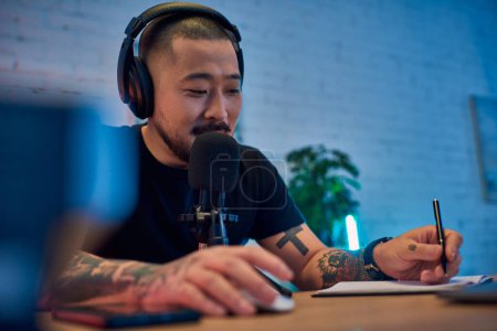 A handsome Asian man wearing headphones speaks into a microphone in his podcast studio.