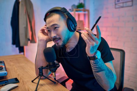 Un joven asiático graba un podcast en su estudio, usando auriculares y hablando en un micrófono.