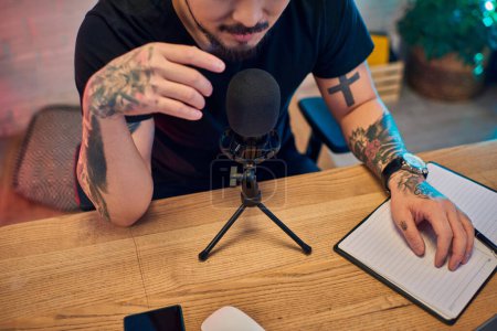 A tattooed Asian man recording a podcast in his studio.