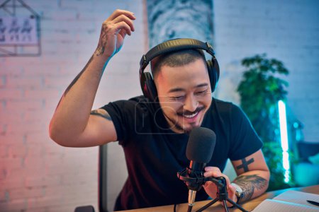 Un Asiatique souriant enregistre son podcast dans un home studio, portant des écouteurs et parlant dans un micro.