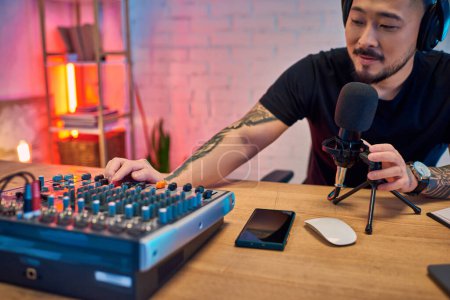 An Asian man records a podcast in his home studio, using a microphone and audio mixer.