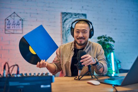 A handsome Asian man smiles while holding a vinyl record, podcasting in his studio.
