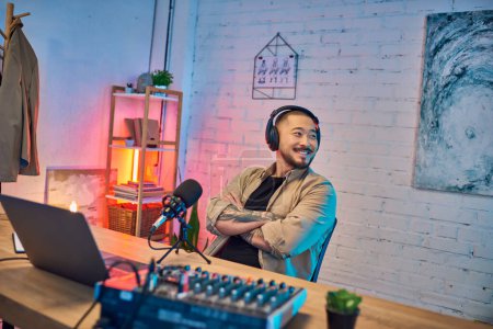 An Asian man smiles brightly while wearing headphones and recording in his home podcast studio.
