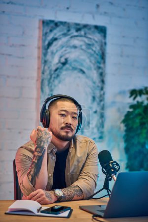 Un Asiatique tatoué avec un casque est assis dans son studio, regardant attentivement la caméra tout en enregistrant un podcast.