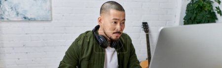 A handsome Asian man, wearing headphones, focuses intently on a computer screen in his studio, surrounded by instruments.