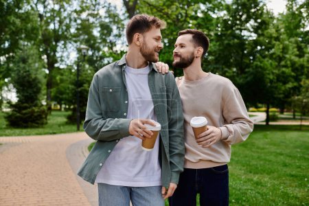 Two bearded men, dressed casually, stroll hand-in-hand through a green park, enjoying a shared moment and a cup of coffee.