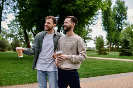 Two bearded men, dressed casually, share a laugh while walking in a park, each holding a cup of coffee.