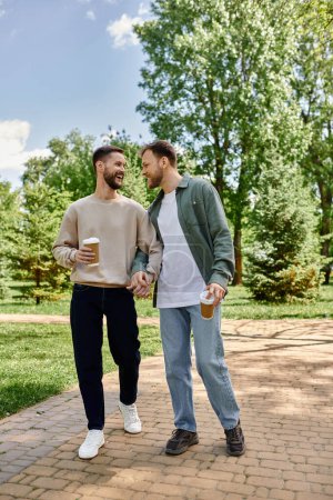 A happy, bearded gay couple walks hand-in-hand through a lush green park on a sunny day.