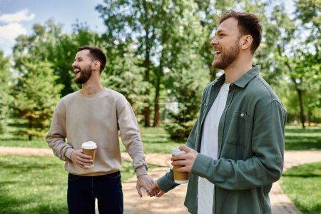 Two bearded men in casual clothes walk hand-in-hand in a park, smiling and enjoying each others company.