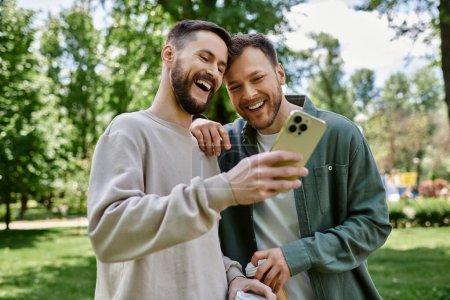 Two bearded gay men laugh together while looking at a smartphone in a green park.