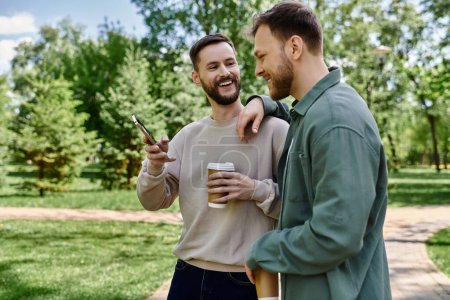 Two bearded men in casual clothing laugh together in a green park, enjoying a sunny day.