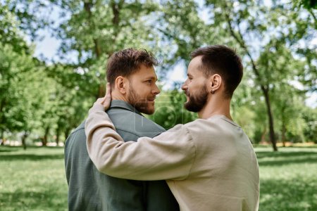 A bearded gay couple stands in a verdant park, embraced and looking into each others eyes.
