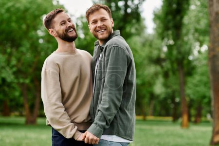 Two bearded men, an LGBTQ couple, laugh together in a green park on a sunny day.
