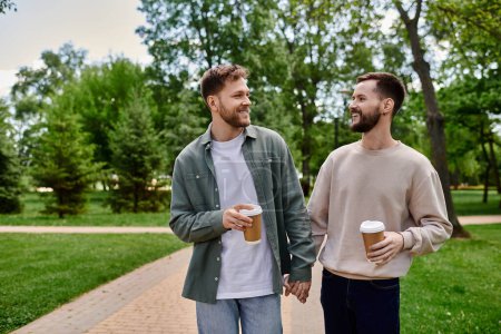 A bearded gay couple walks hand-in-hand through a park, enjoying a coffee together.