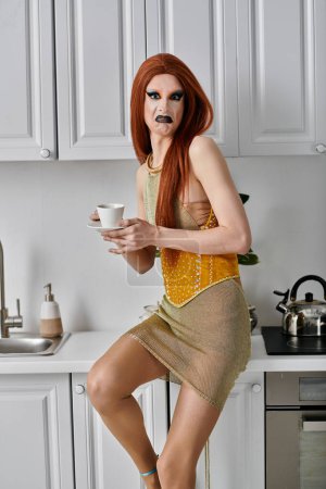 A drag queen in a sparkling gold dress enjoys a cup of coffee in a modern kitchen.