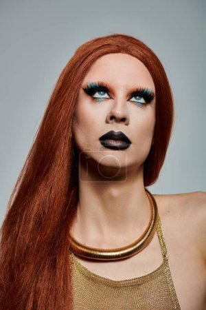 A drag queen with long red hair and dramatic makeup, wearing a gold top and necklace.
