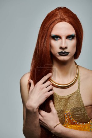 Foto de A drag performer with long red hair, blue eyeshadow, and a golden necklace and top poses for a photo shoot. - Imagen libre de derechos