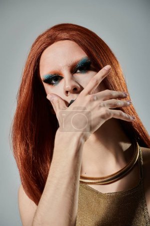 Foto de A drag queen with red hair and dramatic makeup poses, hand over mouth, wearing a gold sequined top and choker necklace. - Imagen libre de derechos