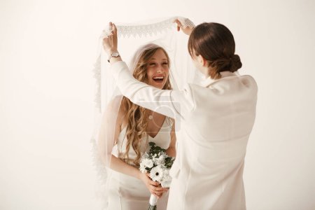 Two brides in white wedding attire share a joyful moment during their wedding ceremony, as one playfully unveils the others veil.