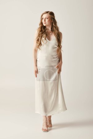 A young bride stands in a flowing white slip dress against a white background.