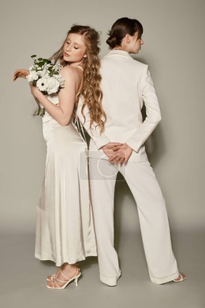 A lesbian couple in white wedding attire poses against a grey background, celebrating their love.
