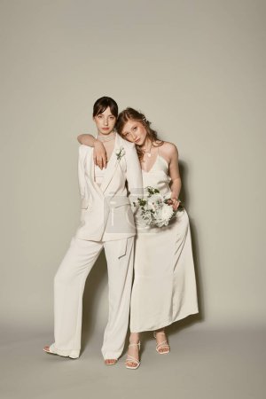 Zwei Frauen in weißer Hochzeitskleidung stehen zusammen, eine hält einen Strauß weißer Blumen in der Hand und feiert ihre Liebe vor grauem Hintergrund..