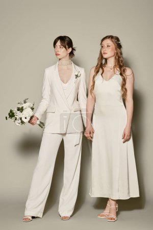 Zwei junge Frauen in weißen Gewändern stehen bei ihrer Trauung vor grauem Hintergrund zusammen.