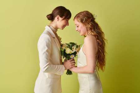 Una joven pareja lesbiana, vestida de blanco, se sonríe mientras sostiene un ramo de flores durante la ceremonia de su boda..