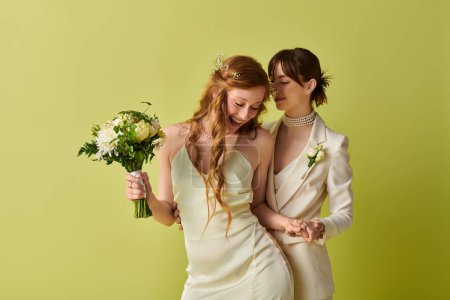 Two women in white wedding attire laugh together, holding a bouquet of white flowers.