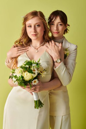 Dos mujeres en traje de novia blanco abrazan, con uno sosteniendo un ramo de flores, sobre un fondo verde.