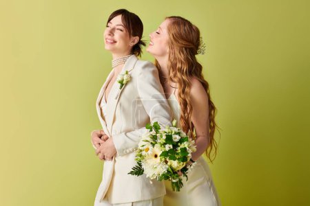 Two brides in white attire stand back-to-back, smiling and holding a bouquet of flowers against a green background.