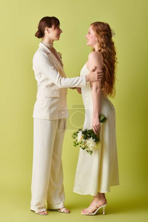 Two brides in white wedding attire stand facing each other, smiles on their faces, on a green background.