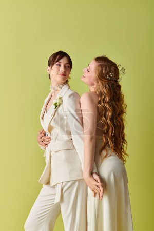Two women in white wedding attire stand side-by-side, smiling and looking at each other against a green background.