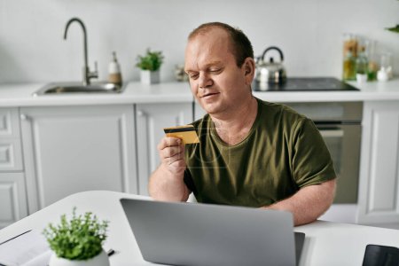 Ein inklusiver Mann sitzt an einem Küchentisch und betrachtet eine Kreditkarte, während er einen Laptop benutzt.
