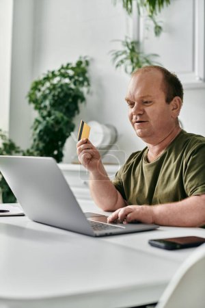 Un hombre con inclusividad se sienta en una mesa con un portátil y una tarjeta de crédito en la mano.
