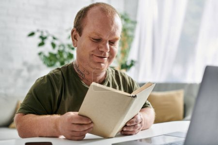 Un hombre con inclusividad se sienta en una mesa en su casa, absorto en un libro. Está vestido casualmente con una camisa verde..