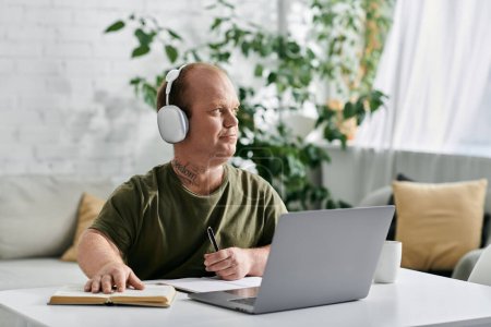 Un hombre con inclusividad usando auriculares se sienta en un escritorio con una computadora portátil, un libro y un bolígrafo, trabajando desde casa.