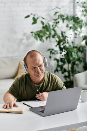 Un hombre con inclusividad en atuendo casual, con auriculares, se sienta en un escritorio con una computadora portátil y libro abierto, concentrándose en su trabajo.