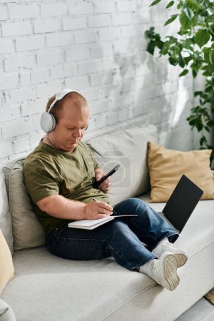 A man with inclusivity wearing headphones sits comfortably on a couch, taking notes in a notebook.