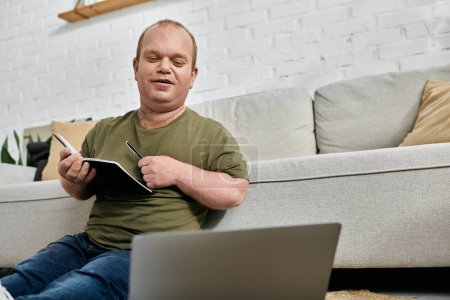 Un hombre con inclusividad se sienta en un sofá en su casa, trabajando en un portátil y tomando notas en un cuaderno.