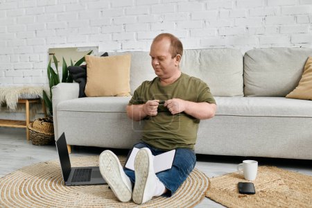 A man with inclusivity sits on the floor in a living room, working on a laptop. He is wearing casual clothes and has a cup of coffee nearby.