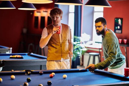 Friends enjoy a casual game of billiards, one taking a shot while the other watches and holds a drink.