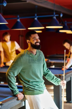 Ein Mann mit Bart lehnt an einem Billardtisch und lächelt seine Freunde beim Billardspielen an.