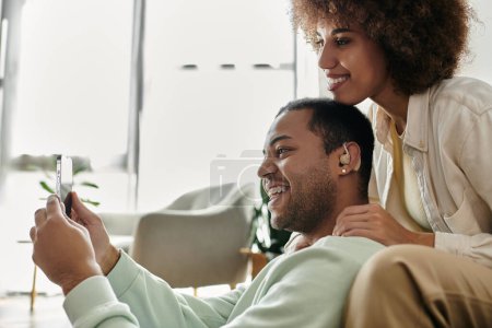 Joyeuse femme afro-américaine assise près de son mari avec un appareil auditif et regardant le téléphone.