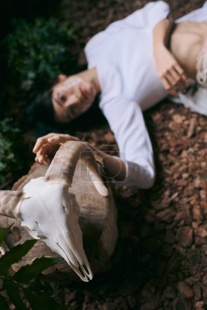 Eine Frau liegt in einem sumpfigen Wald, ihre Hand ruht auf einem weißen Schädel.