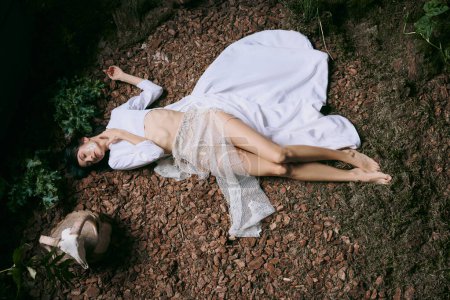 A woman in a white dress lies on ground and foliage.