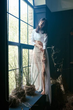 A woman in a white top and sheer skirt stands near a window with green plants, gazing out.