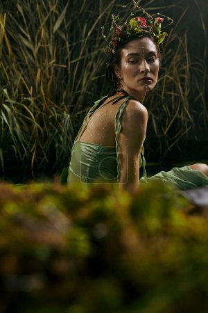 A woman wearing a green dress and a flower crown poses in a swamp.